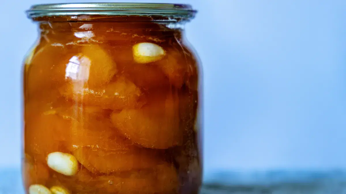A close-up of a jar filled with preserved garlic and peaches, set against a soft blue background.